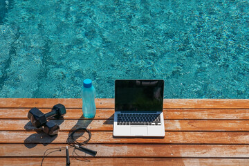 Fitness concept image of a dumbbell, a jump rope, a bottle of water and a computer for online workouts near the pool. Close up, outside.