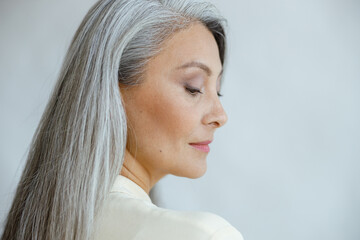 Pretty middle aged Asian woman with loose straight grey hair poses on light background in studio...