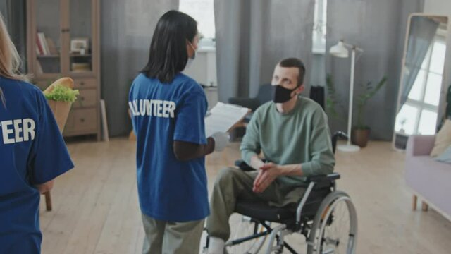 Tracking rear-view slowmo of team of volunteers in blue T-shirts and protective face masks bringing food and clothes in cardboard boxes helping young man with disability in wheelchair