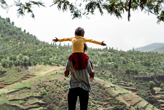 black mother holding on shoulders black little daughter on top of mountain