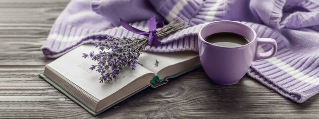 Morning coffee. A cup of coffee on a wooden table, an open book and a warm sweater against the background of a bouquet of flowers. Still life in lavender color. Cozy morning. © Tetiana
