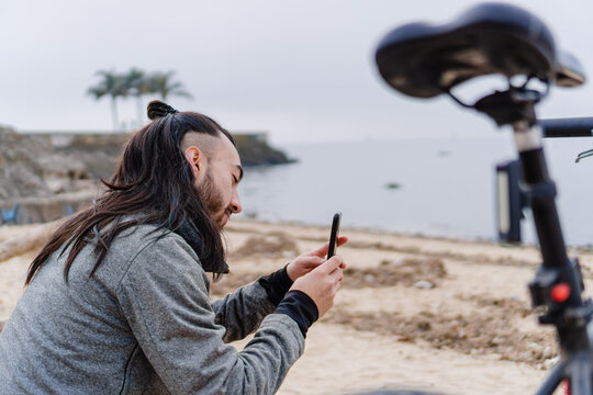 young latino male long-haired hispanic cyclist sportsman taking a picture with his smart phone for his social networks, sitting on the beach in the morning.