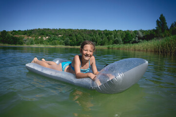 A little cute girl in a swimsuit swims on an inflatable mattress on a blue career lake.