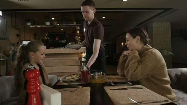 a man waiter brings food to his mother and daughter sitting in a cafe. Family lunch. High quality FullHD footage