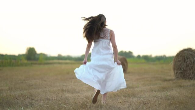 Beautiful pregnant woman running in wheat field with haystacks at summer day