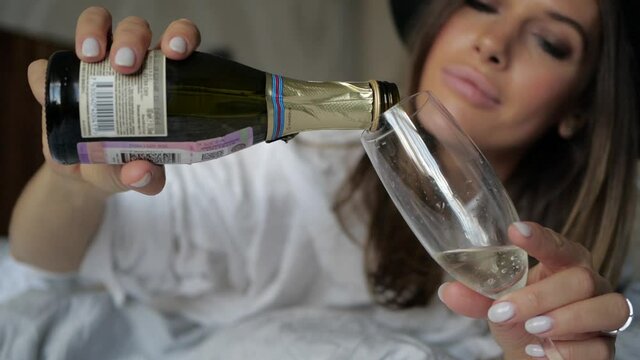 cheerful young woman pours champagne from a bottle into a glass and drinks it looking directly into the camera. High quality FullHD footage