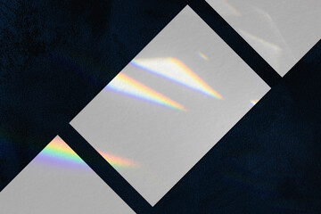Three empty white horizontal rectangle a4 poster, business card mockups lying diagonally with overlay of rainbow light refraction caustic effect and shadow on trendy dark blue concrete background.