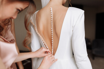 Bridesmaid's hands helping bride fastens with buttons on the back of a beautiful white wedding lace...