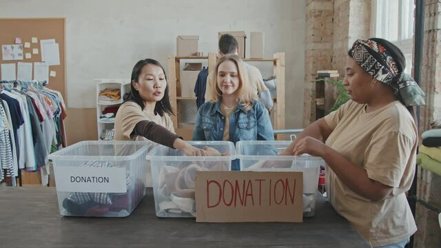 Medium slowmo shot of three multi-ethnic women from volunteer organization standing by table, sorting out donations from plastic containers, while men carrying heavy cardboard boxes