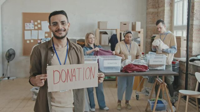 Medium slowmo shot of smiling Middle-Eastern man holding cardboard sign calling for donations while his coworkers from volunteer organization sorting out donated clothes