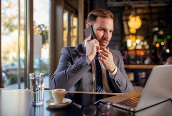 Caucasian businessman sitting at table in cafe, talking on mobile phone, drinking coffee and typing on laptop