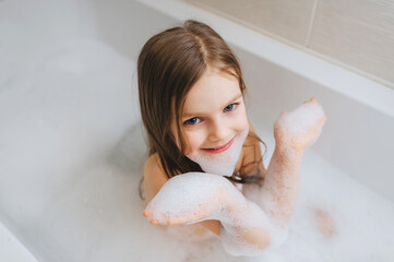 A small, smiling, beautiful red-haired girl with long hair, a child bathes, bathes in a white bath...