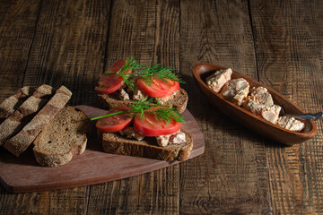 On a wooden background, there are ready-made sandwiches with cod liver, tomato and greens, canned fish are lined in a dark plate.