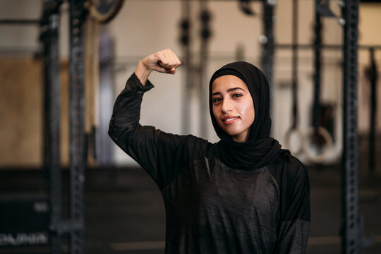 Smiling fit Muslim sportswoman showing biceps in gym