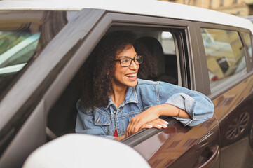 Happy young smiling African American woman black haired driver in glasses sitting in new brown car, smiling looking at camera enjoying journey. Driving courses and life insurance concept.