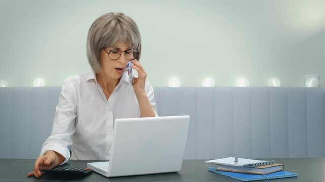 An elderly gray-haired angry businesswoman indignantly communicates with her subordinate employees on the phone and solves business issues online on the phone and laptop, sitting at home. The concept