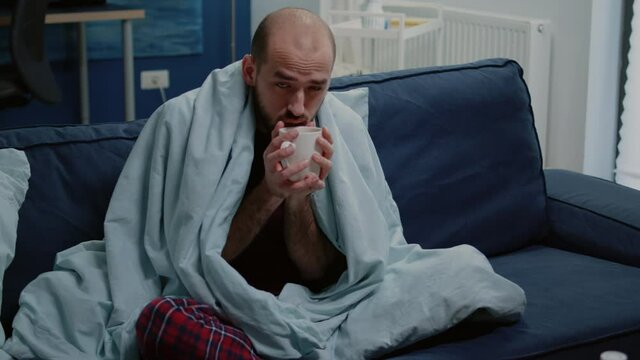 Man with cold shivering and drinking cup of tea while wrapped in blanket. Sick person holding mug with hot drink against chills and fever symptoms while looking at camera. Adult with flu