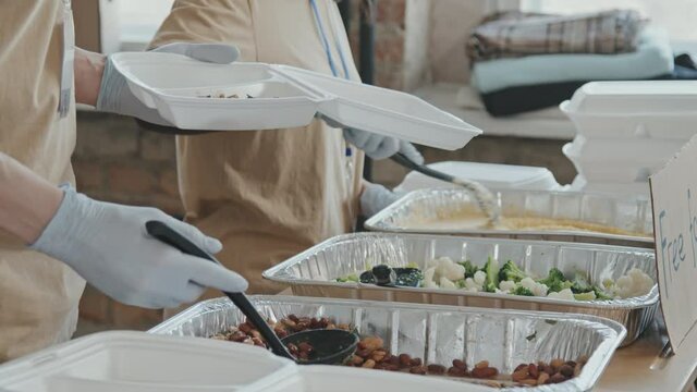 Midsection shot of team of volunteers putting food in plastic containers ang giving it to homeless and needy people
