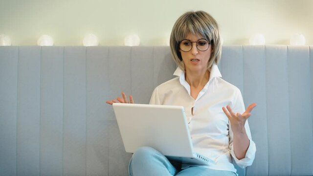 An elderly gray-haired angry businesswoman indignantly communicates with her subordinate employees on the phone and solves business issues online on the phone and laptop, sitting at home. The concept