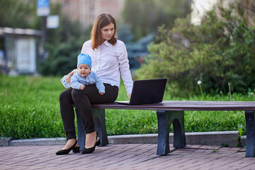 Young mother with baby in her knees remote work online using laptop with an internet connection outdoors, sitting on bench in park.