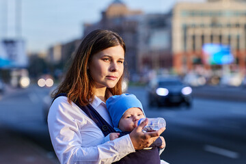 Young woman gives drink to baby in sling on street.