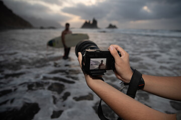 Close-up of the hands of a photographer holding his camera during a photo session with a surfer model at the beach during sunset - Photoshoot concept.