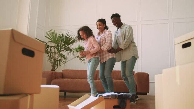Happy african american family of three dancing with domestic plant among lots of cardboard boxes at new own apartment