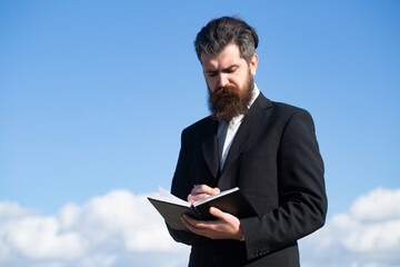 Business man with sky. Portrait with copy space of serious man holding copybook and with pen with thoughtful expression over sky background.