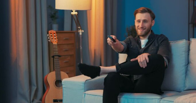 A young man is relaxing on the sofa in the living room in the evening, he is dressed in black, in his hand he is holding the TV remote control, he is watching a TV series, a program, a match