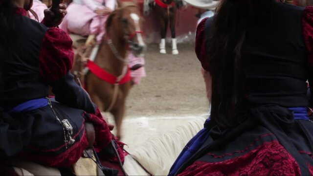 Two mexican escaramuzas or skirmish watching their team riding horses in choreographed synchronized maneuvers to music performance, taking photo and video with a mobile phone