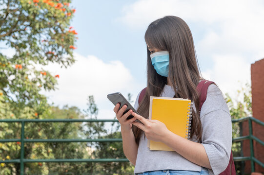 University Student Wearing Protective Mask, Holding A Notebook And Using A Smartphone