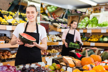 Fototapeta premium Young happy positive smiling woman seller holding half of watermelon in hands in fruit store