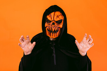 Man dressed in pumpkin latex mask and hooded velvet cape, scares with his hands, on orange background. Halloween and days of the dead concept.