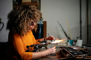Side view of a woman lampworker working with glass tubes and torch in her workshop