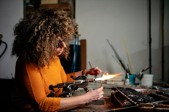 Side view of a woman lampworker working with glass tubes and torch in her workshop