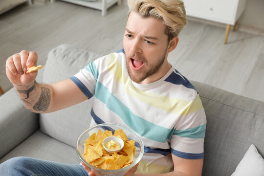 Handsome young man eating tasty nachos while watching TV at home