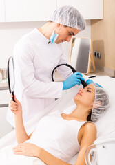 Young woman getting needle-free mesotherapy for face and looking in mirror during procedure