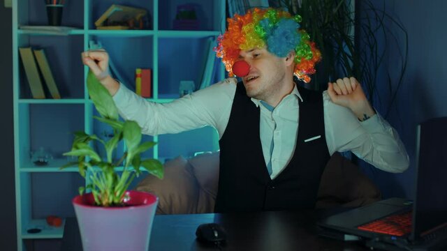 A young man in the image of a clown dances in the workplace, sitting at a computer in the office. The concept of a clown at work