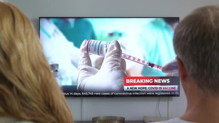 Family Couple Watching TV News Sitting on Couch in Living Room Together. TV Presenter Telling Breaking News about Coronavirus, Lockdown and Vaccinations. Rear View with Dolly-in