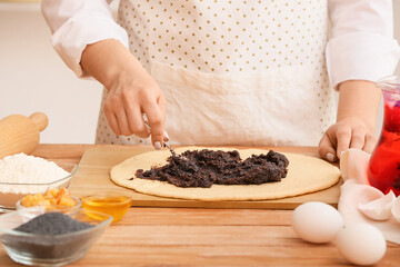 Woman preparing tasty poppy seed bun at table in kitchen, closeup