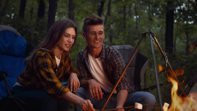 Camping and picnic concept. Young affectionate man and woman frying marshmallow on fire, enjoying evening in forest