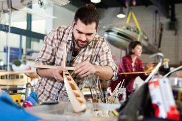 Young glad cheerful positive smiling handsome bearded man enjoying his hobby - modeling light airplanes in aircraft hangar