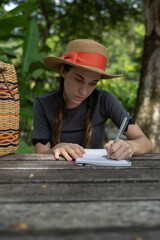 Caucasian Girl wearing a straw hat sitting in the park on a wooden bench writes in her diary or...