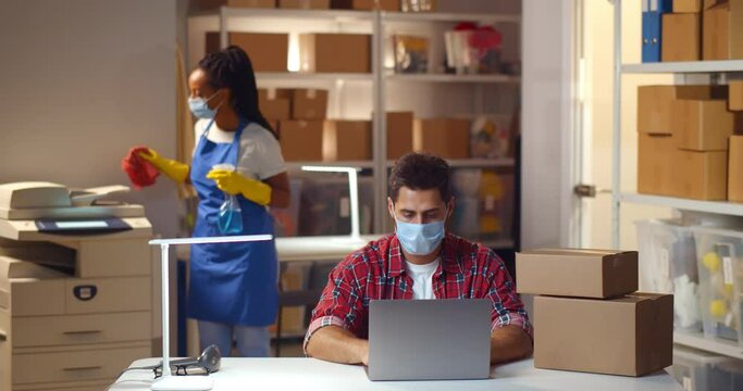 Small business owner in mask working on laptop in warehouse with maid cleaning on background