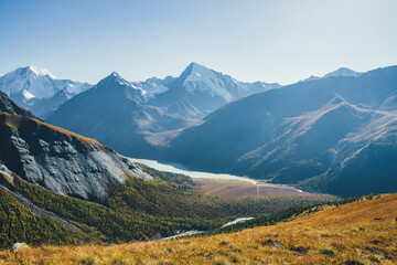 Fototapeta premium Wonderful alpine landscape with mountain lake and mountain river in valley with forest in autumn colors on background of snowy mountains silhouettes under blue sky. Beautiful mountain valley in autumn