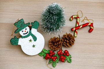 Top view studio shot of Christmas eve festival decorative items smiley face snowman with green hat gloves and scarf Small mock up tree Pine seeds Cherry fruit and Candy cane sticks on wooden table