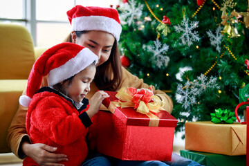 Fototapeta na wymiar Happy Asian family daughter girl wears sweater red and white Santa Claus hat sitting with mom unboxing open present gift box celebrating Xmas eve near Christmas pine tree in living room at home