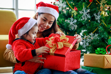 Happy Asian family daughter girl wears sweater red and white Santa Claus hat sitting with mom unboxing open present gift box celebrating Xmas eve near Christmas pine tree in living room at home