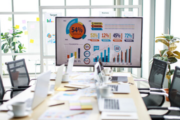 Laptop computers and white hot coffee cups on small dish placed on working table full of company paper report documents in meeting room in front of target growth rate graph and chart on big screen