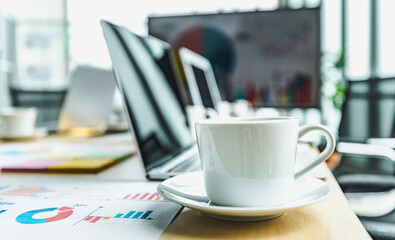 White hot tasty coffee cup on small dish placed on working table with laptop computer in meeting room in front target growth rate graph and chart report on big screen monitor in blurred background